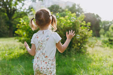 Back view little caucasian happy kid girl 5-6 years old in white casual clothes blow bubbles play on park green sunshine lawn, spending time outdoor in village countyside during summer time vacations.