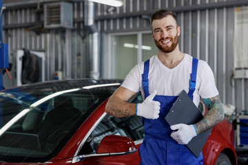 Young smiling male professional car mechanic man in denim blue overalls white t-shirt gloves hold clipboard with papers document show thumb up work in light modern vehicle repair shop workshop indoor