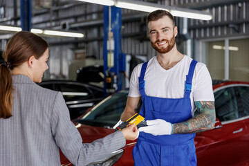 Young smiling happy professional car mechanic man in blue overalls gloves hold payment terminal fow paying with credit card by female owner driver woman work in vehicle repair shop workshop indoors