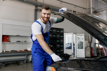 Strong smiling happy troubleshooter young male professional technician car mechanic man in denim blue overalls white t-shirt raised hood bonnet work in light modern vehicle repair shop workshop inside