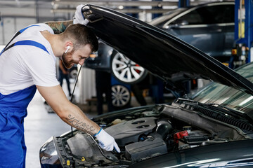 Obraz na płótnie Canvas Young professional technician car mechanic man in blue overalls white t-shirt gloves listen music in earphones fix problem with raised hood bonnet work in modern vehicle repair shop workshop indoor.