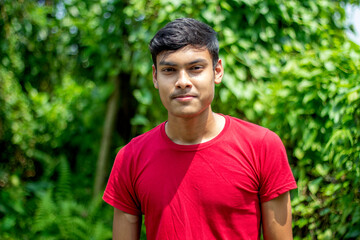 A young man in a red T-shirt is standing look at the camera. he has a fair complexion and beautiful lips and the background behind him is blurry