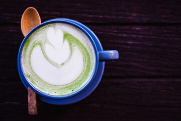 The blue beautiful ceramic cup of hot matcha milk tea which have latte art as a flower with wooden spoon on the surface of the latte on the dark brown wooden table background with coyspace