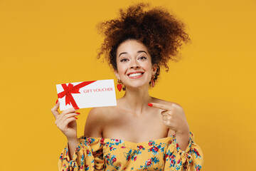 Young happy satisfied smiling woman 20s with culry hair wear casual clothes hold point finger on gift certificate coupon voucher card for store isolated on plain yellow background studio portrait. - obrazy, fototapety, plakaty