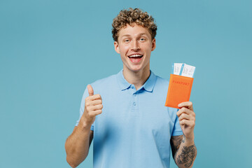 Traveler tourist satisfied young curly man 20s years old wear azure t-shirt hold passport boarding tickets showing thumb up like gesture isolated on plain pastel light blue background studio portrait