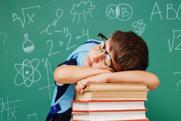 Young tired male kid school boy 5-6 years old in t-shirt backpack glasses lying sleep on books isolated on green wall chalk blackboard background. Childhood children kids education lifestyle concept