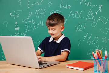 Young smiling fun male kid school boy 5-6 years old in t-shirt sitting use laptop pc cocmputer isolated on green wall chalk blackboard background Childhood children kids education lifestyle concept