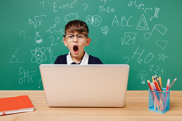 Young male kid school boy 5-6 years old in t-shirt glasses sit use laptop pc cocmputer shouting isolated on green wall chalk blackboard background Childhood children kids education lifestyle concept