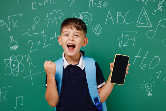 Young male kid school boy 5-6 years old in t-shirt backpack use mobile cell phone blank screen workspace do winner gesture isolated on green wall chalk blackboard background. Childhood kids concept