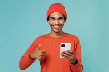 Young smiling happy african american man in orange shirt hat use hold mobile cell phone show thumb up gesture isolated on plain pastel light blue background studio portrait People lifestyle concept