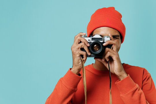 Young smiling happy african american man 20s in orange shirt hat taking photo picture on retro vintage photo camera isolated on plain pastel light blue background studio. People lifestyle concept