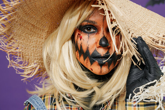 Close up young creepy woman with Halloween makeup mask in scarecrow costume cover painted face with straw hat brim isolated on plain dark purple background studio Celebration holiday party concept.