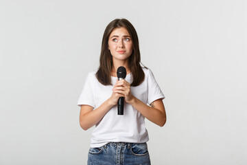 Shy cute brunette girl scared of singing in public, standing with microphone and looking nervous