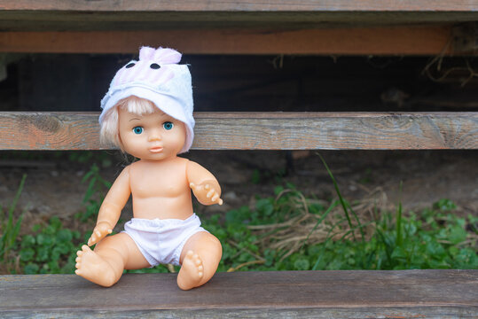 Cute kewpie doll in hat and shorts sits on rustic wooden bench in country house yard space for text. Vintage children toy