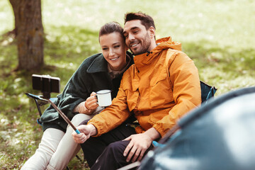 camping, tourism and travel concept - happy couple with smartphone drinking tea and taking selfie at tent camp