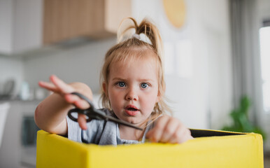 Portrait of little cauccasian girl using scissors indoors at home, looking at camera.