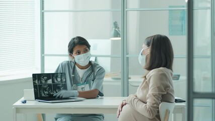 Hispanic midwife in protective mask and gloves showing ultrasound scan on laptop to Caucasian pregnant woman during prenatal visit in time of covid-19 pandemic