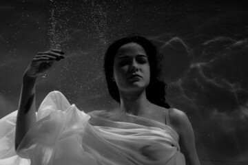 Black and white photo where a beautiful girl in a white dress swims underwater in the pool, and she...