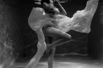 Black and white photo where a beautiful girl in a white dress swims underwater in the pool, and she looks like a siren or a mermaid