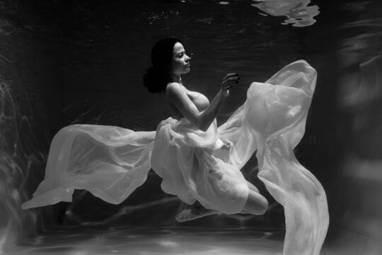 Black and white photo where a beautiful girl in a white dress swims underwater in the pool, and she looks like a siren or a mermaid