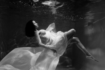 Black and white photo where a beautiful girl in a white dress swims underwater in the pool, and she...