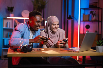 Excited multi ethnic couple with joysticks in hands looking at computer screen while playing video...