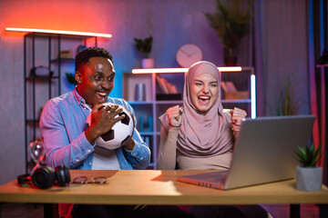 Overjoyed muslim woman and african man gesturing and screaming while watching football match on...