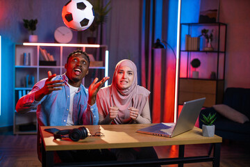 Fototapeta na wymiar African american man throwing up soccer ball while muslim woman in hijab looking at camera with worried facial expression. Young couple spending evening time at home for watching football game.