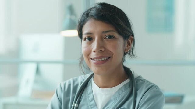 Chest up portrait shot of beautiful Hispanic female doctor in medical uniform with stethoscope over her neck looking at camera and happily smiling