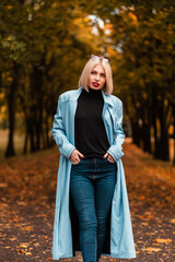 Pretty blonde woman with red lips in fashion blue coat, black sweater and jeans walks in the autumn park. Fall female style and beauty