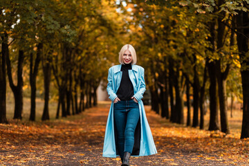 Happy businesswoman in fashion clothes with coat, sweater and jeans walks in autumn park with fall leaves