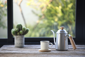 White coffee cup and stainless coffee drip brew pot with cactus on table in front of window
