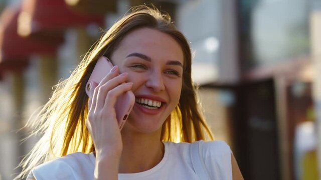 Happy woman with long blonde hair wearing white t-shirt talking on phone on street, parallax background. Arc shot young female in city at sunset. Concept of communication