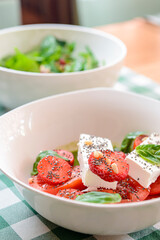 Caprese salad with strawberry, mozzarells, basil and balsamic sauce served in a white bowl over green plaid background.