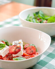 Caprese salad with strawberry, mozzarells, basil and balsamic sauce served in a white bowl over green plaid background.