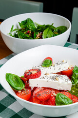 Caprese salad with strawberry, mozzarells, basil and balsamic sauce served in a white bowl over green plaid background.