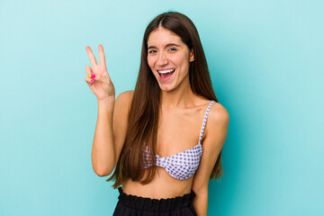 Young caucasian woman wearing bikini isolated on blue background joyful and carefree showing a peace symbol with fingers.