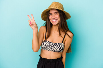 Obraz na płótnie Canvas Young caucasian woman going to the beach wearing bikini isolated on blue background joyful and carefree showing a peace symbol with fingers.