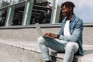 black african american man typing on a laptop on the street in front of a glass building sitting, online work away