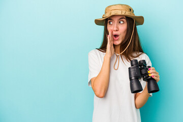 Young caucasian explorer woman holding binoculars isolated on blue background is saying a secret...
