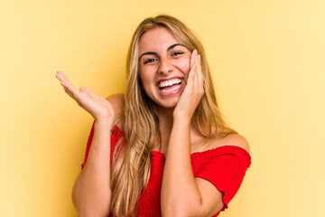 Fototapeta premium Young caucasian blonde woman isolated on yellow background holds copy space on a palm, keep hand over cheek. Amazed and delighted.