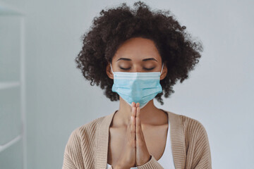 Beautiful young African woman in protective face mask praying with his hands clasped and eyes closed