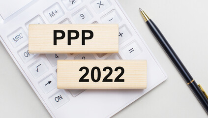 Wooden blocks with the text PPP 2022 lie on a light background on a white calculator. Nearby is a black handle. Business concept