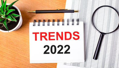 On a wooden table there are reports, a potted plant, a magnifying glass, a black pen and a notebook with the text TRENDS 2022. Business concept