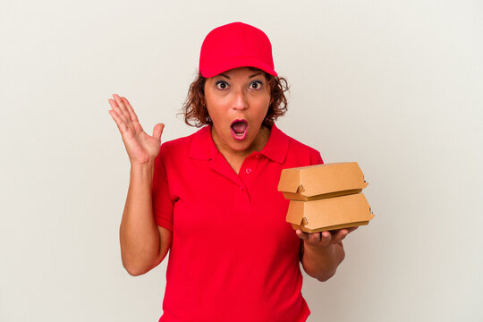 Middle age delivery woman taking burguers isolated on white background surprised and shocked.