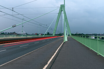 Empty walkway and bicycle lane along a road over a suspension bridge on a cloudy summer evening. A tramway runs in the middle of the road. Light trails left by passing cars..