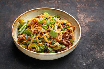 Close up of oriental noodles in a bowl, on black stony background. Beef fried on a wok with rice noodles, green beans, zucchini, pak choi and green peppers in spicy soy-oyster sauce. Thai cousine dish