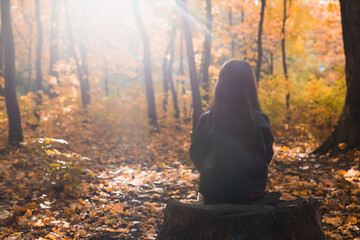 Child girl sits alone on a wooden stump while walking through the forest on an autumn day....