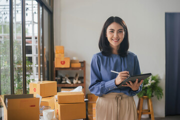 Portrait business Asian woman smiling look at camera and use tablet checking information on parcel shipping box before send to customer. Entrepreneur small business working at home.