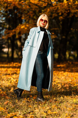 Pretty young stylish woman in vintage sunglasses with an elegant blue coat walks in an autumn park with yellow autumn foliage on a sunny day